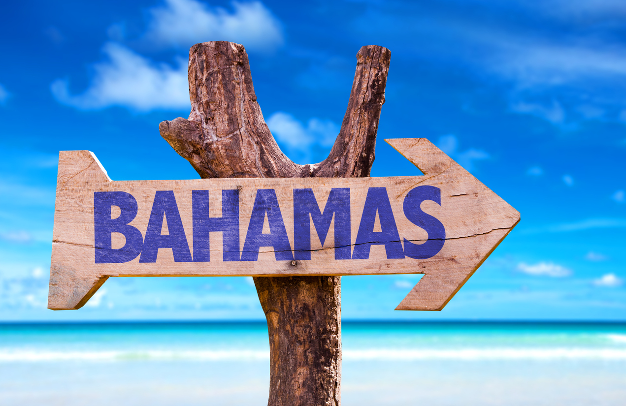 blue skies and sand of a beach with a wooden sign in the foreground attached to a tree. The sign is in the shape of an arrow and reads BAHAMAS.